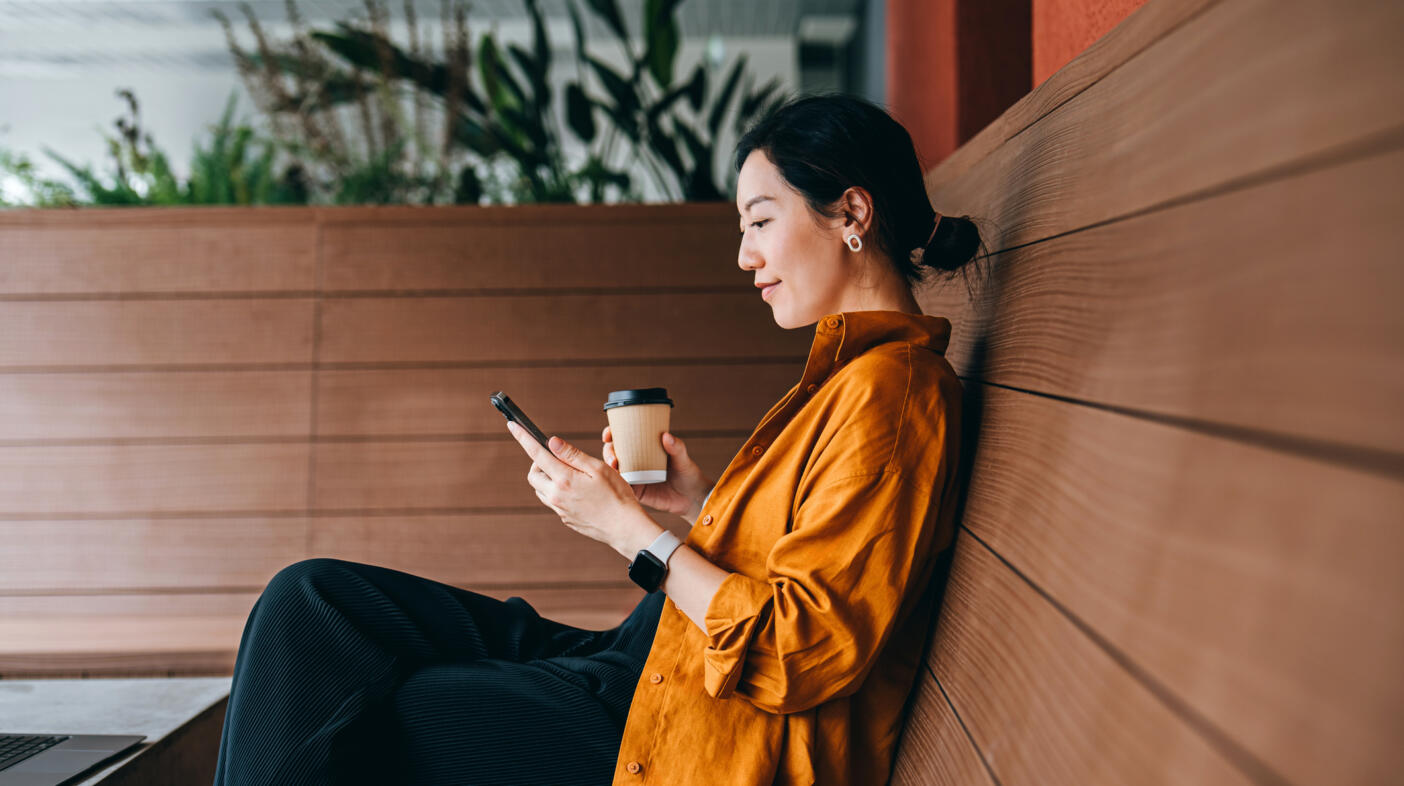 Young Asian woman sitting in a sidewalk cafe with a cup of coffee and using her smartphone. Enjoying a relaxing moment in the afternoon.