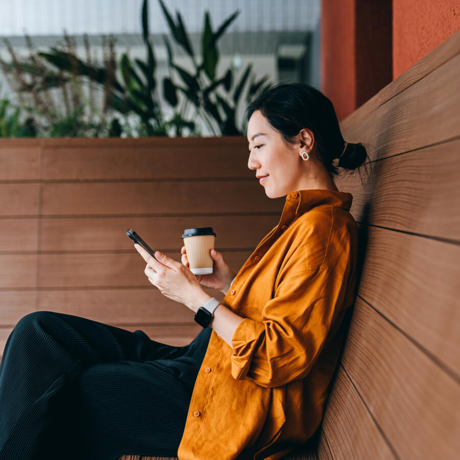 Young Asian woman sitting in a sidewalk cafe with a cup of coffee and using her smartphone. Enjoying a relaxing moment in the afternoon.